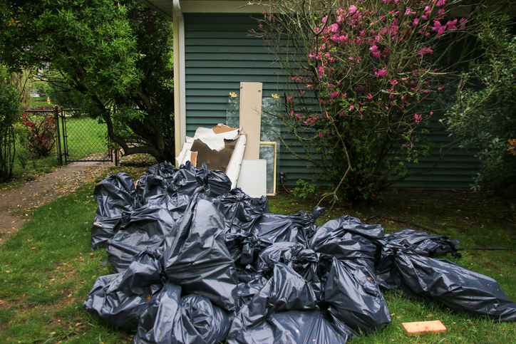 Black trash bags are stacked in a yard, surrounded by trees and blooming flowers.